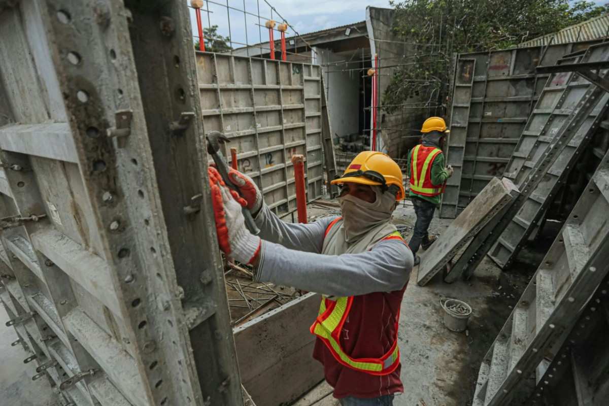 Two construction workers, one in the foreground hammering a metal formwork panel, are seen on a building site.